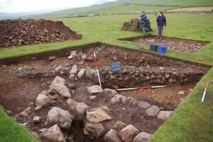 Archeologische opgraving in een grasveld met meetstokken en stenen